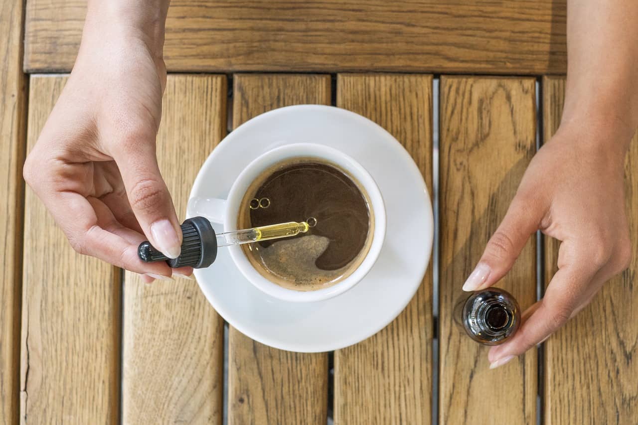 A person adding CBD oil from a dropper into a cup of coffee on a wooden table.