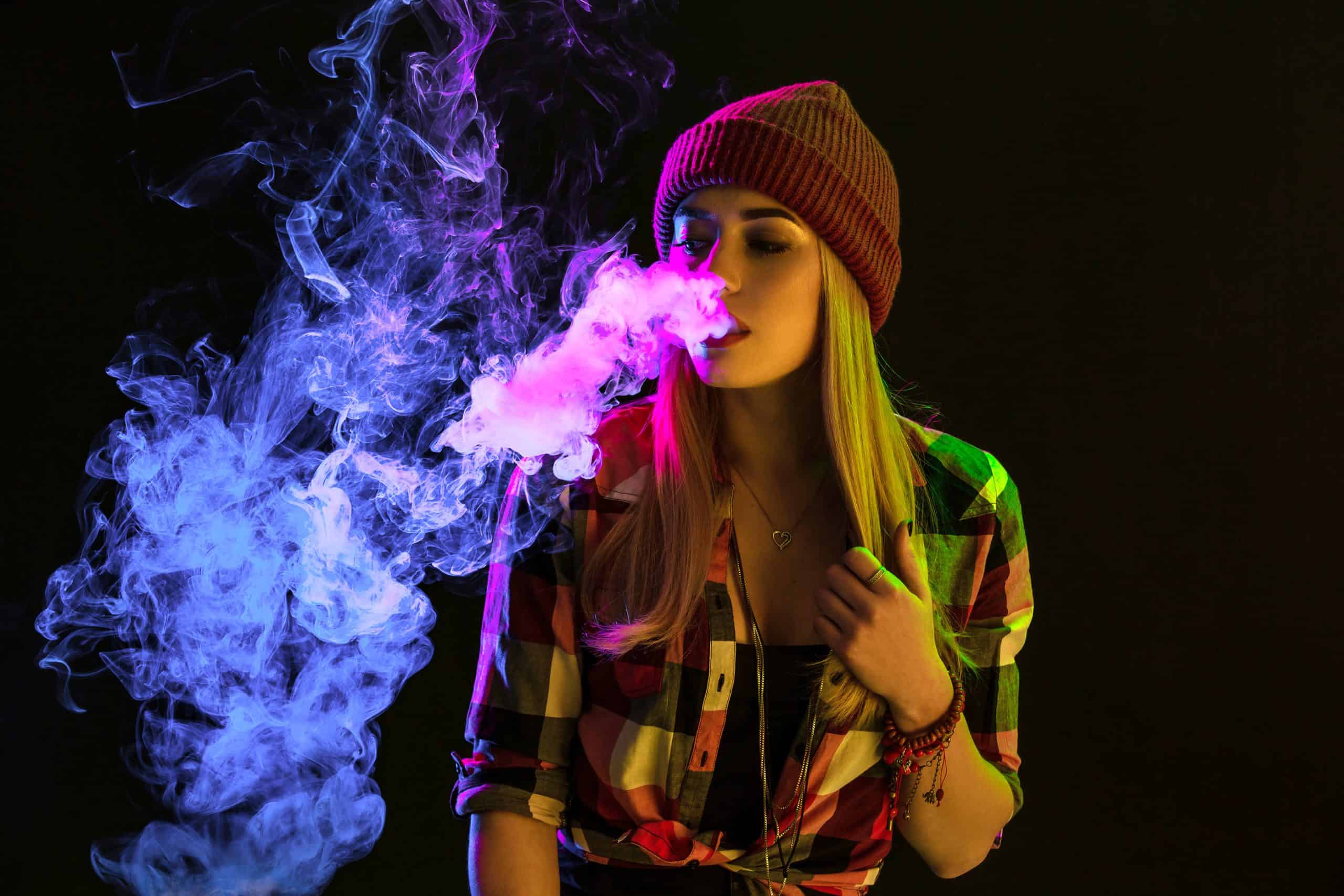 A person exhaling colorful vape smoke while wearing a beanie and plaid shirt.