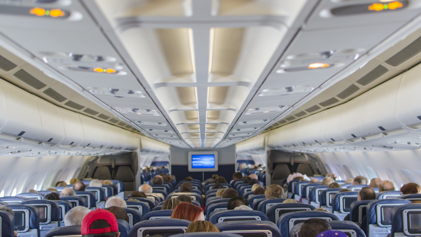 Interior view of an airplane cabin filled with passengers, showing overhead compartments and a screen.